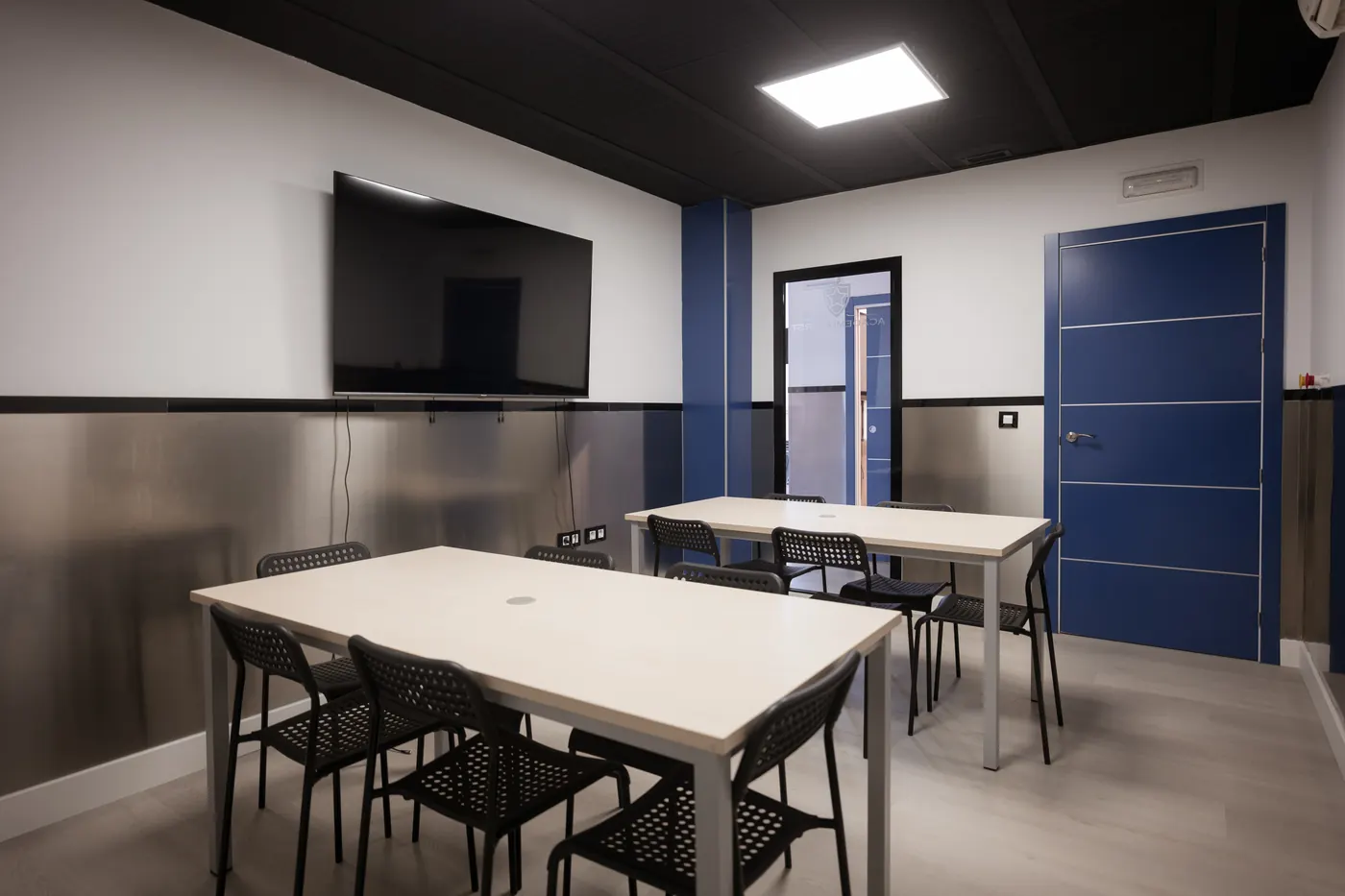 Aula renovada con dos mesas, pantalla y puerta azul.