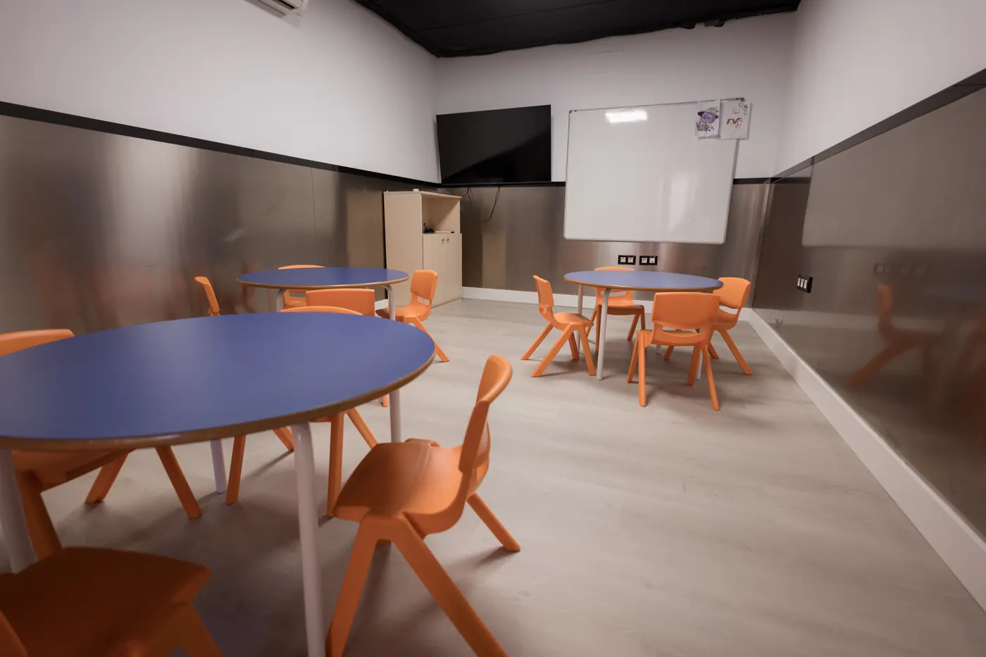 Aula de infantil con mesas redondas azules y sillas naranjas.