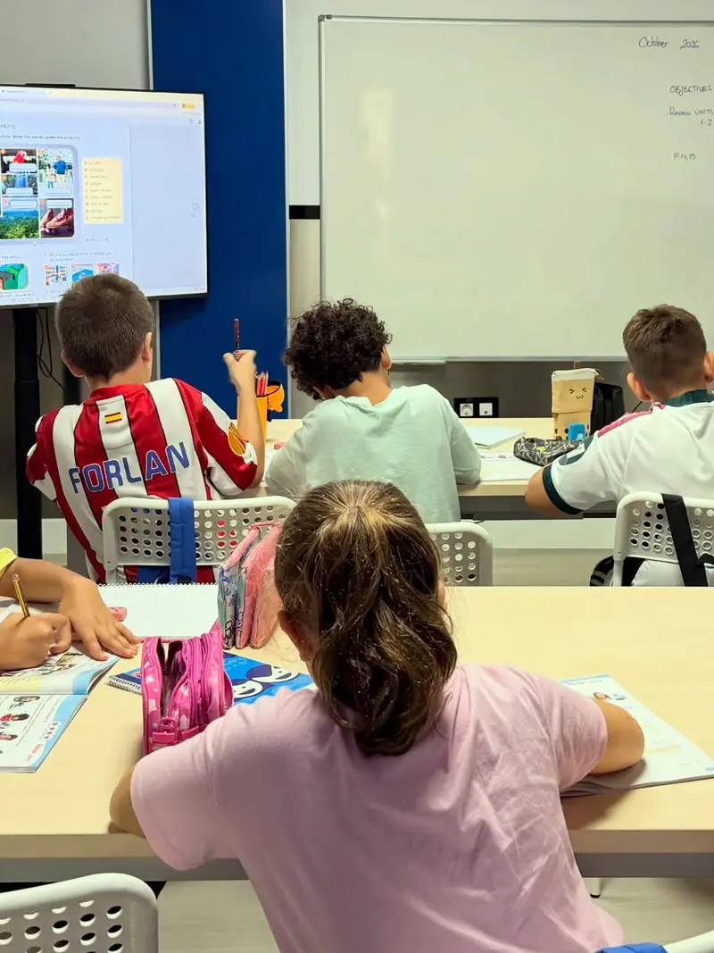 Clase vista desde atrás: cuatro alumnos de espaldas trabajan con sus libros frente a una pantalla con un ejercicio de inglés y una pizarra blanca con los objetivos del día.