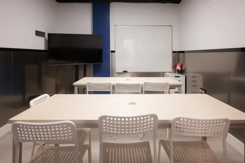 Aula de First vista desde una mesa larga con sillas blancas; al fondo, una pantalla y una pizarra blanca.