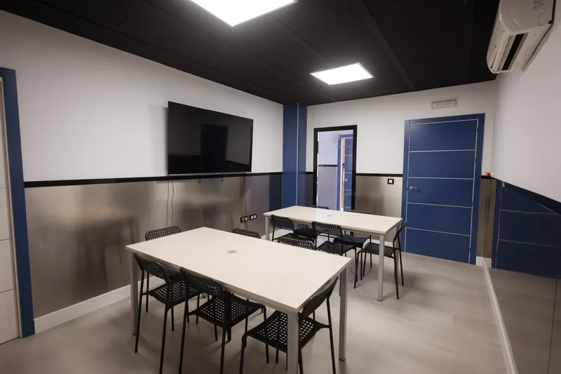 Aula de First con dos mesas largas de madera clara y sillas negras, una pantalla en la pared y una puerta azul.