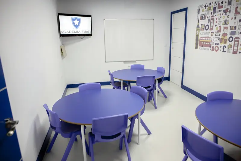 Aula con mesas redondas y sillas de tamaño infantil, una pizarra blanca y una pantalla en la pared.