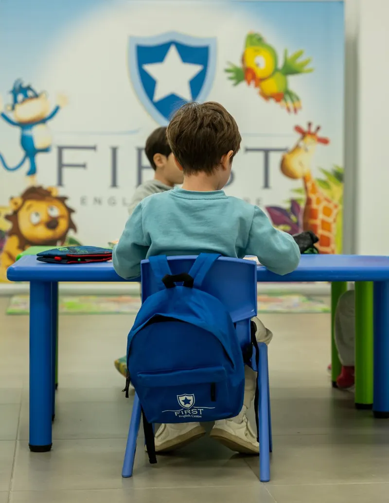 Alumno de First English Centre con la mochila del centro en el aula de Pre-Starters, de espaldas, frente al cartel con el escudo y las mascotas.
