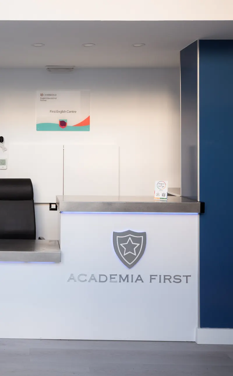 Recepción del centro: mostrador blanco con el escudo de First, una silla negra y una pared azul.