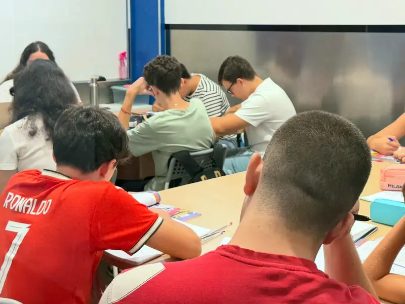 Alumnos de secundaria escriben alrededor de una mesa larga, vistos desde atrás.